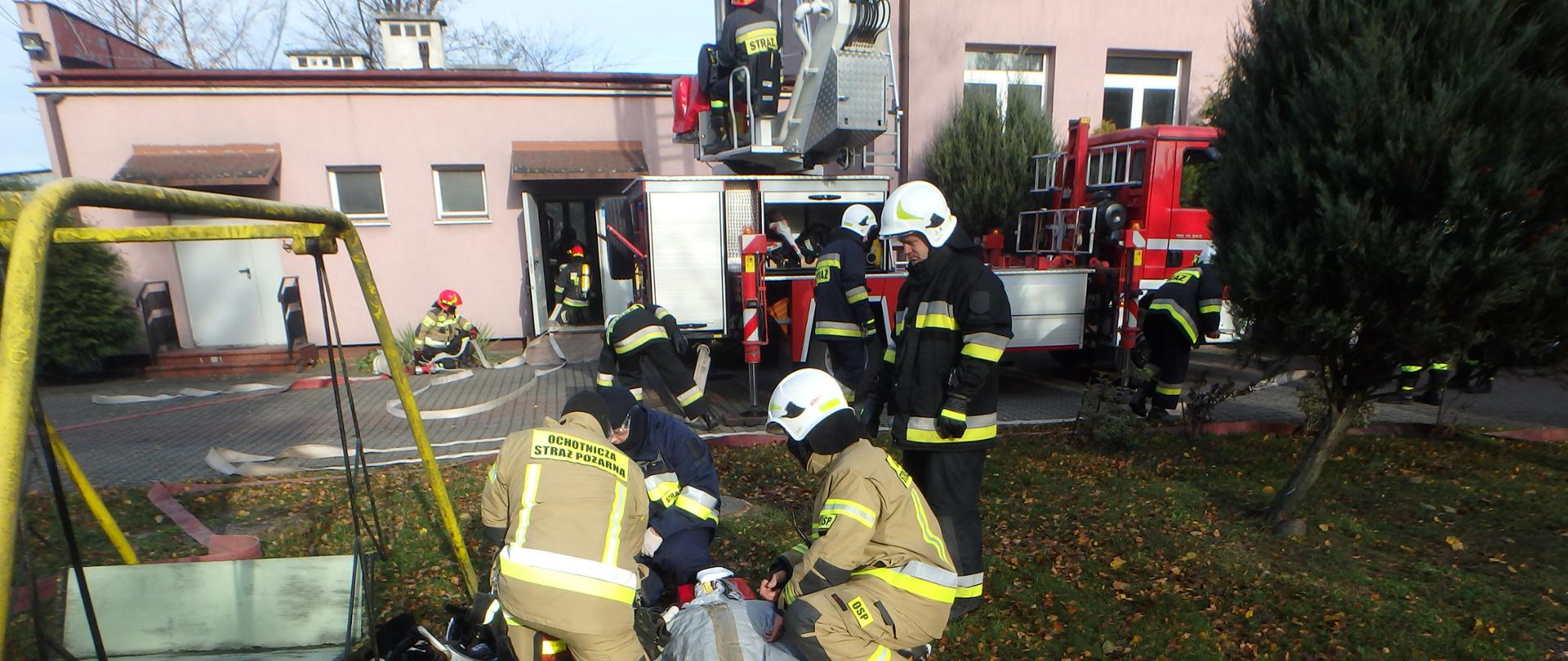 Przed budynkiem DPSu stoi rozłożony, czerwony samochód ratowniczo-gaśniczy. Obok samochodu chodzą strażacy w umundurowaniu specjalnym. Na ziemi leżą rozłożone węże strażackie. Na pierwszym planie widać manekina leżącego na desce ortopedycznej. Obok manekina są strażacy, którzy w ramach ćwiczeń udzielają mu pomocy medycznej.