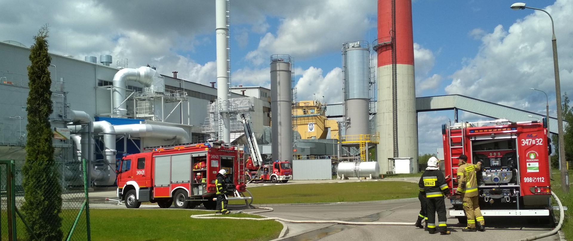 Na tle zakładu widać samochody pożarnicze zasilające się z hydrantów zewnętrznych i podające wodę na plac. Dodatkowo przy instalacji przy budynku sprawiono drabinę mechaniczna z koszem.