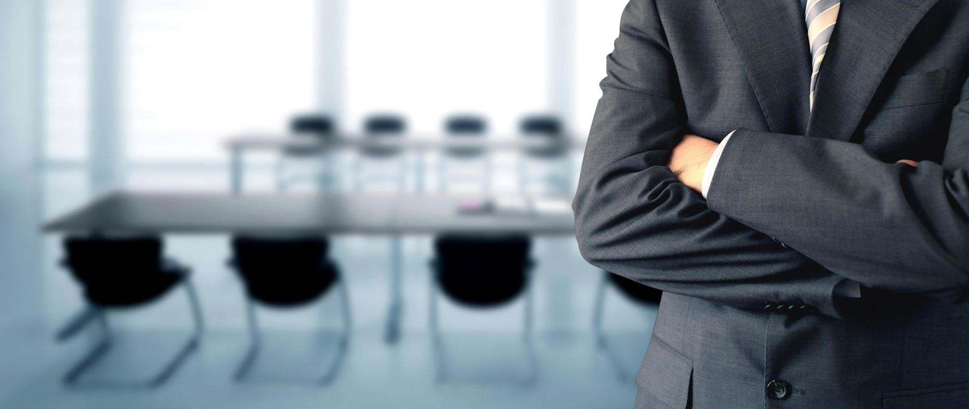Mężczyzna w garniturze z założonymi rękami. W tle sala konferencyjna.