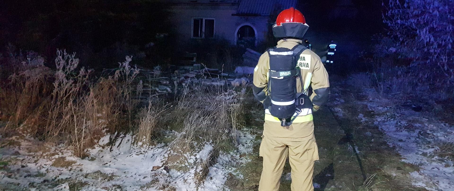 Dom jednorodzinny w którym doszło do pożaru nocną porą. Na pierwszym planie strażak wyposażony w aparat powietrzny. 