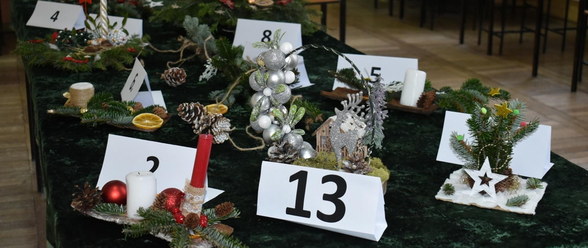 Widok na stół przykryty zielonym obrusem, na którym ustawiono kilkanaście różnych stroików bożonarodzeniowych z numerami startowymi.