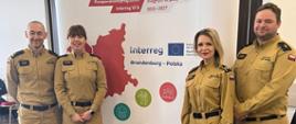 Czworo funkcjonariuszy Państwowej Straży Pożarnej w mundurach służbowych stoi na tle ścianki promocyjnej programu Interreg Brandenburgia – Polska. Wszyscy uśmiechają się do zdjęcia, prezentując swój udział w projekcie współpracy transgranicznej. Na ściance widoczne logo programu oraz kolorowe elementy graficzne symbolizujące współpracę polsko-niemiecką.