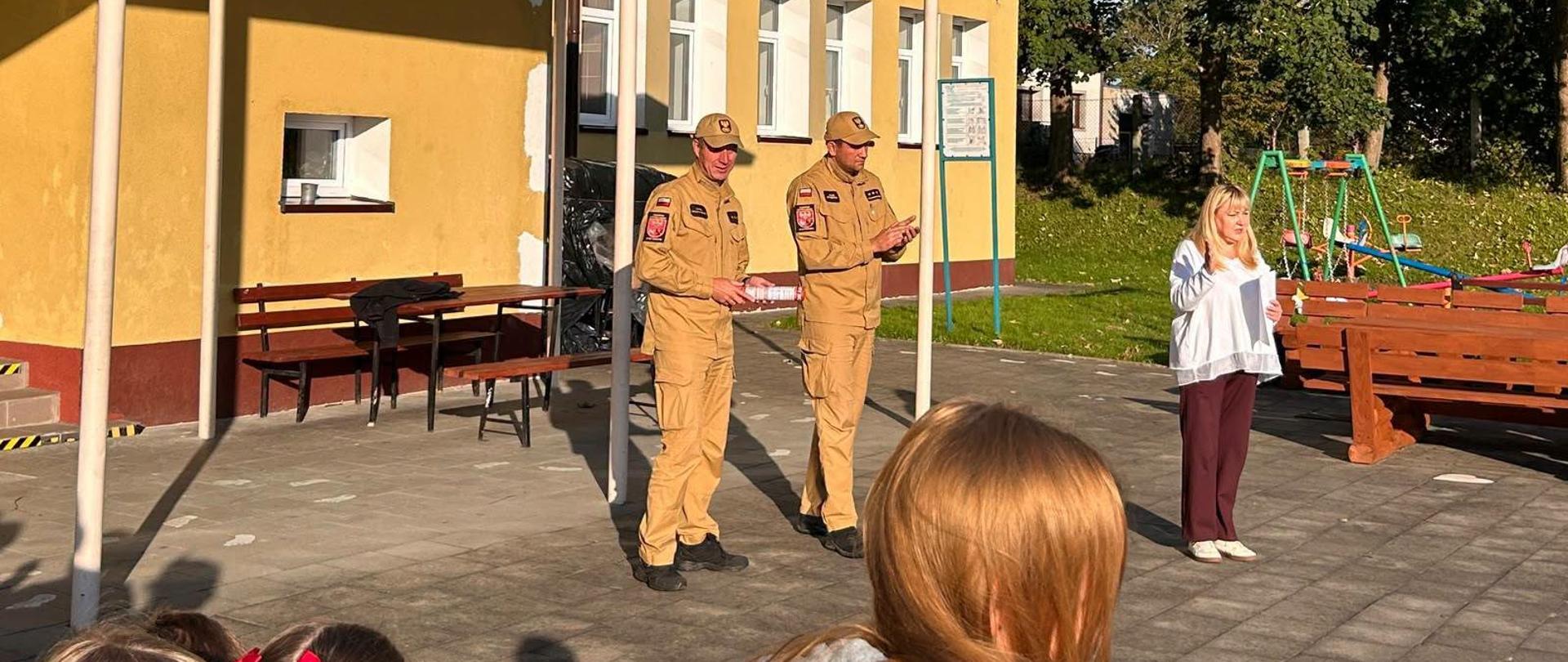 Na fotografii widoczne jest spotkanie strażaków Państwowej Straży Pożarnej z dziećmi i pracownikami placówki oświatowej. Strażacy prowadzą krótką pogadankę dotyczącą zasad bezpieczeństwa oraz właściwego zachowania w sytuacjach zagrożenia. Spotkanie odbywa się na terenie przed budynkiem szkoły i stanowi element działań edukacyjno-profilaktycznych.