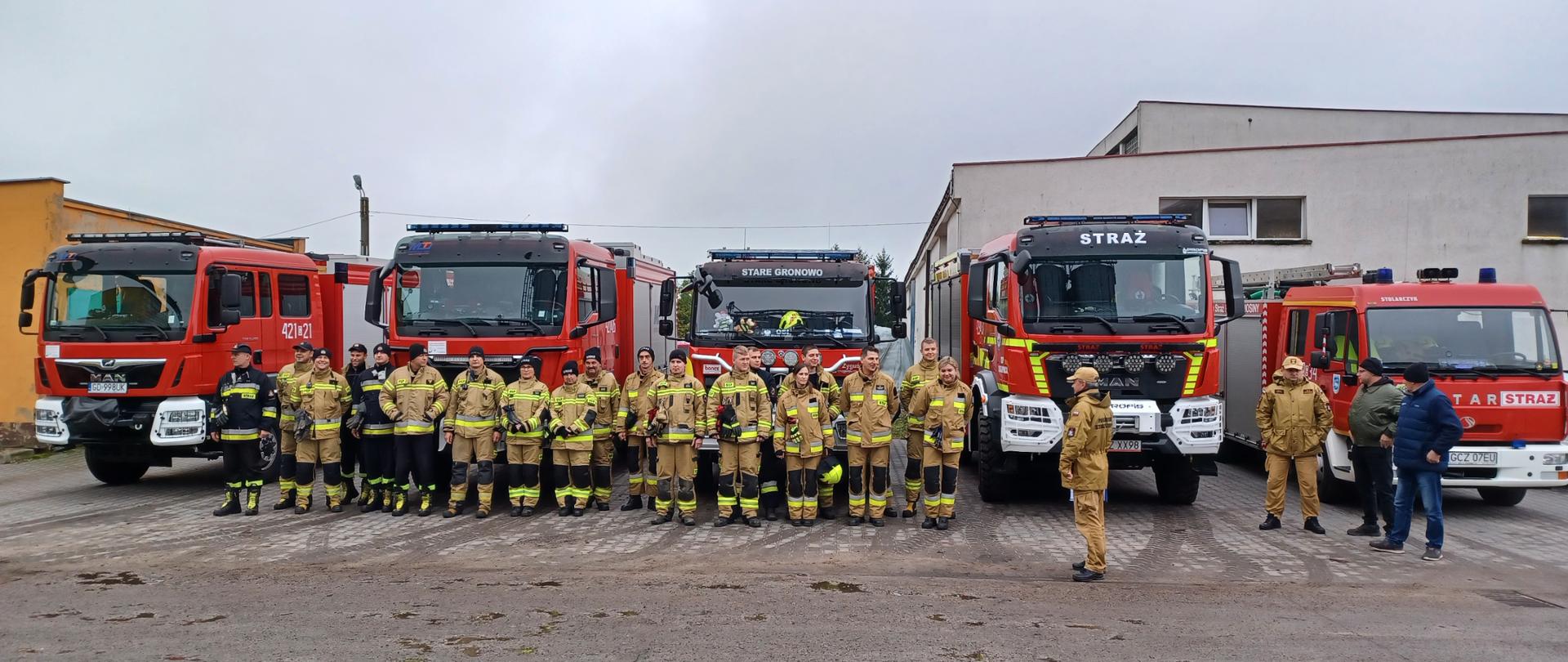 Zdjęcie przedstawia strażaków stojących w dwuszeregu podczas zbiórki na terenie firmy, w tle samochody pożarnicze i budynki.