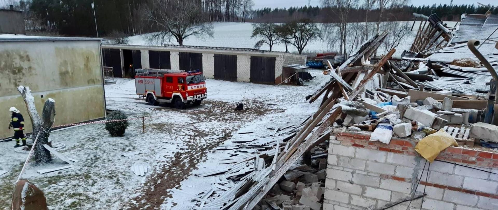 Zdjęcie przedstawia samochód pożarniczy przed zniszczonym budynkiem, widać całkowicie zniszczony dach, połamane elementy leżą na dachu, w tle inne budynki gospodarskie