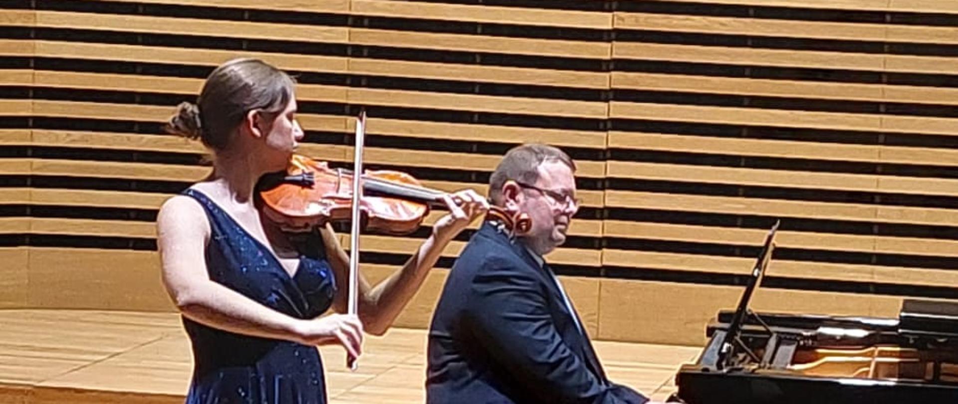 Dziewczyna gra na skrzypcach, mężczyzna akompaniuje na fortepianie.