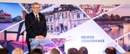 Na podwyższeniu z przodu sali za mównicą stoi wiceminister Murdzek i mówi do mikrofonu, za nim za ścianie napis Bridge Conference.