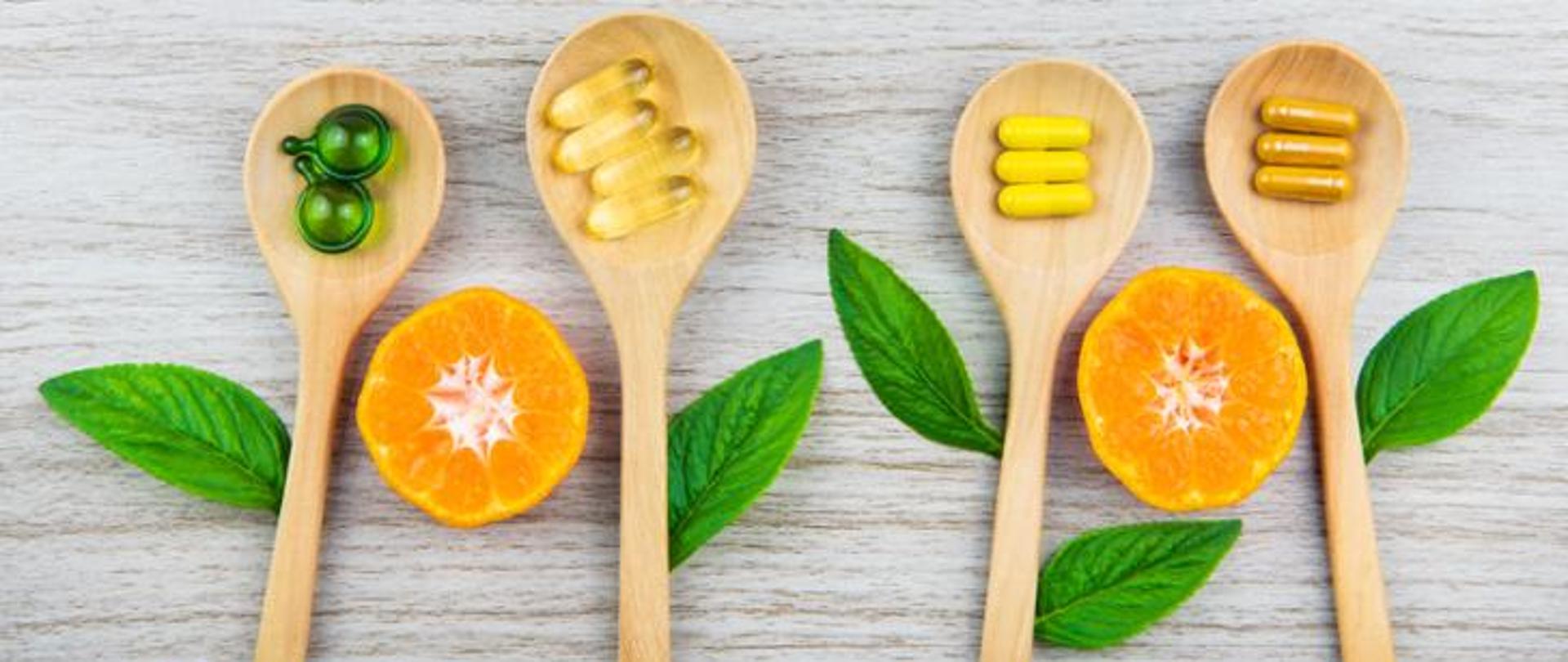 Na zdjęciu znajdują się drewniane łyżki na których ułożone są tabletki, między łyżkami zielone listki mięty i połówki pomarańczy 