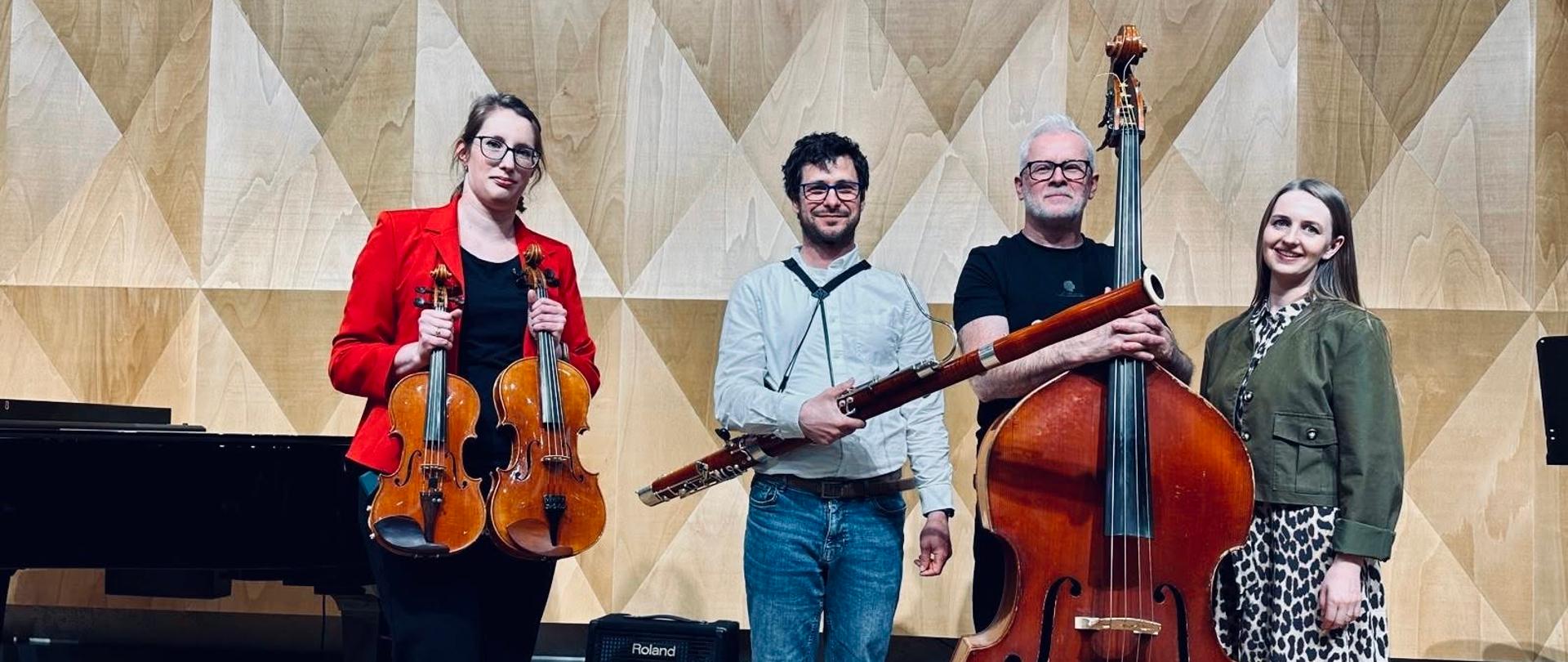 Czworo muzyków stoi w rzędzie na scenie z drewnianą podłogą, na tle geometrycznej ściany z jasnego drewna w kształcie trójkątów. Od lewej: kobieta w czerwonej marynarce trzyma skrzypce, mężczyzna w niebieskiej koszuli trzyma fagot, mężczyzna w czarnej koszulce stoi przy dużym kontrabasie, a obok niego kobieta w sukience w cętki lamparcie. Po lewej stronie widoczny jest fragment czarnego fortepianu.