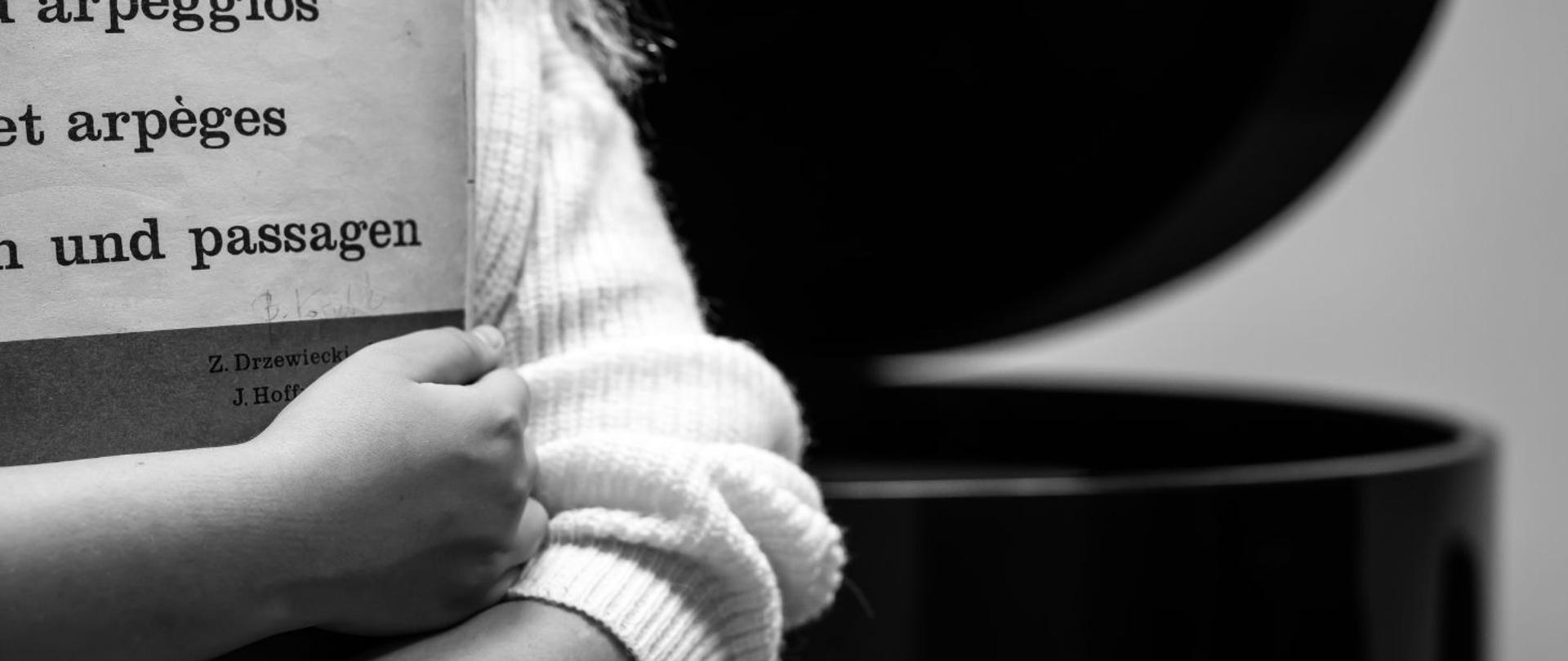 czarno-białe zdjęcie dziewczynki w białym swetrze na tle czarnego fortepianu, dziewczynka obejmuje nuty z napisem “gamy i pasaże”