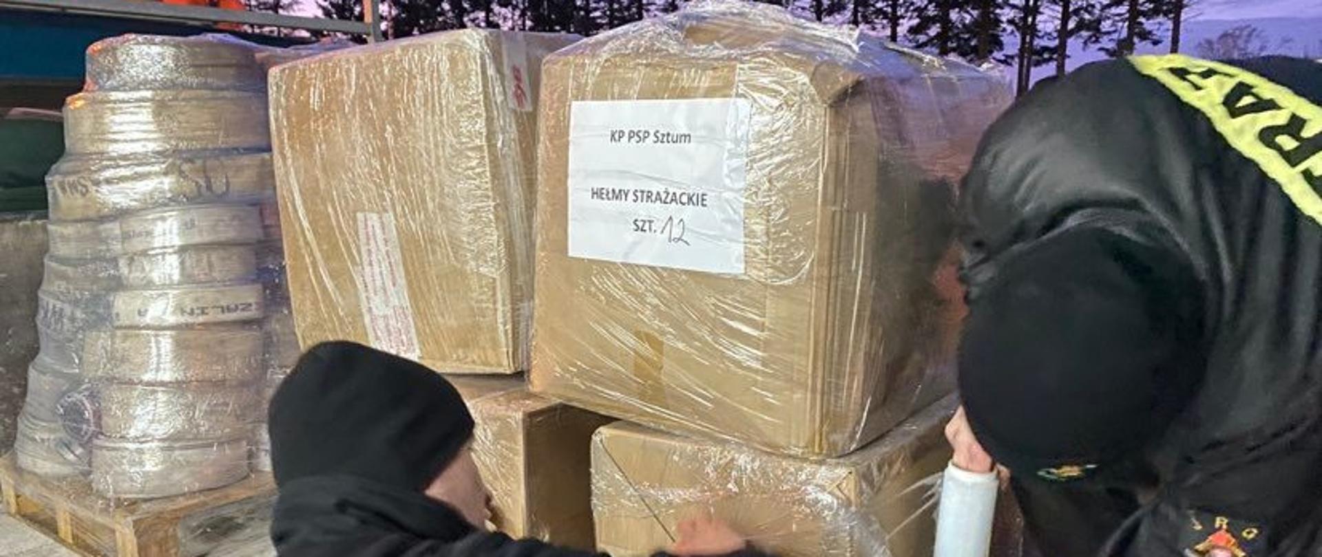 pora nocna, na pace pojazdu ciężarowego strażacy w ubraniach koszarowych owijają kartony stojące na palecie.