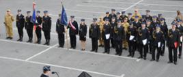 Na zdjęciu widać uroczystą zbiórkę strażacką odbywającą się na placu w Karczewie. W centrum kadru stoją szeregi funkcjonariuszy Państwowej i Ochotniczej Straży Pożarnej w galowych mundurach, tworząc zwartą formację. Na przedzie widoczne są dwa poczty sztandarowe — jeden z biało czerwonym sztandarem z orłem, drugi z niebieskim z napisem „Państwowa Straż Pożarna”. Obok nich stoją strażacy w mundurach polowych oraz kobieta w galowym stroju służbowym.
W tle widać namioty informacyjne z logotypami służb ochrony przeciwpożarowej, biały samochód oznaczony napisem „STRAŻ”, a także czerwony wóz bojowy z numerem 551[26]. Całość odbywa się na otwartym placu miejskim, otoczonym zielenią i budynkami.
Scena przedstawia moment oficjalnych obchodów — prawdopodobnie Dnia Strażaka lub uroczystości wręczenia odznaczeń — w atmosferze powagi, dyscypliny i wspólnoty służby.