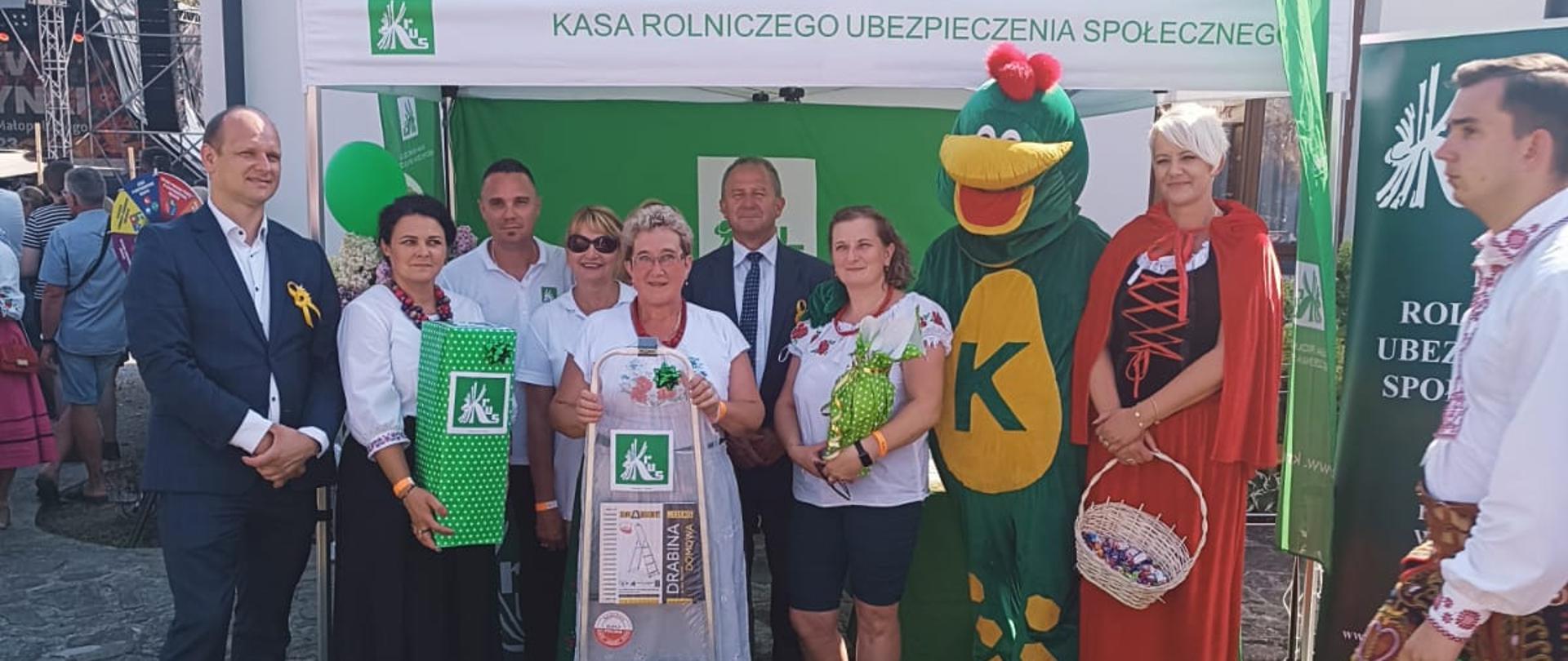 Na tle stoiska informacyjnego KRUS trzech mężczyzn, cztery kobiety – trzy trzymają nagrody zdobyte w konkursie oraz dwie osoby przebrane za Czerwonego Kaptura i smoka Krusika. 