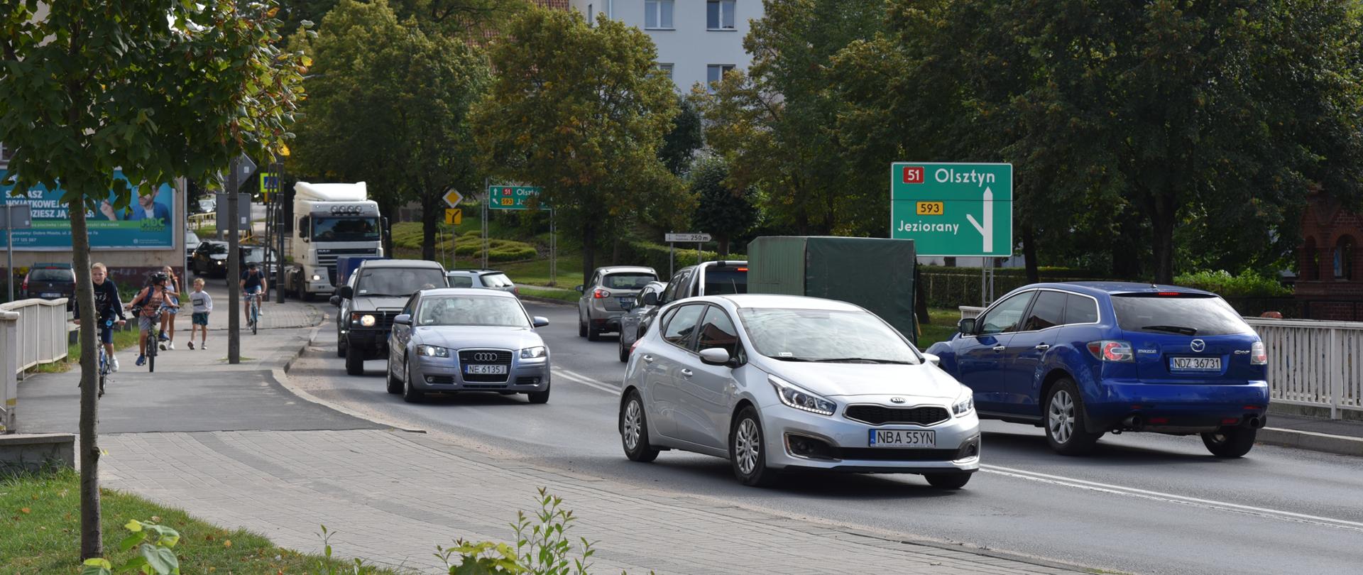 Droga krajowa numer 51 w Dobrym Mieście, z zieloną tablicą informacyjną z kierunkami na Olsztyn i Jeziorany. Na pierwszym planie dwa srebrne auta poruszające się po tej drodze. Po chodniku poruszają się piesi i rowerzyści.