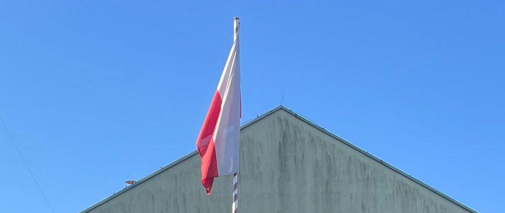 Strażacy podczas zmiany służby, ustawieni w dwóch rzędach oddają honory do wciąganej przez poczet flagowy Flagi Rzeczpospolitej Polskiej. Na środku dowódca zbiórki.