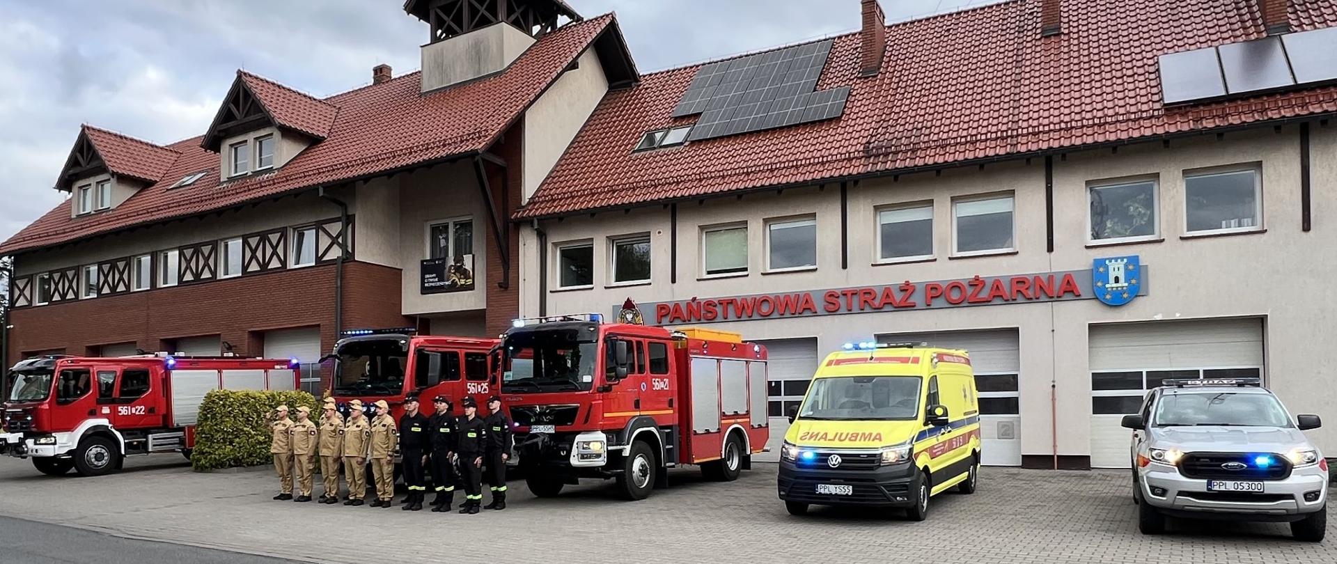 Strażacy oddający hołd poległym funkcjonariuszom. W tle samochody pożarnicze i budynek komendy