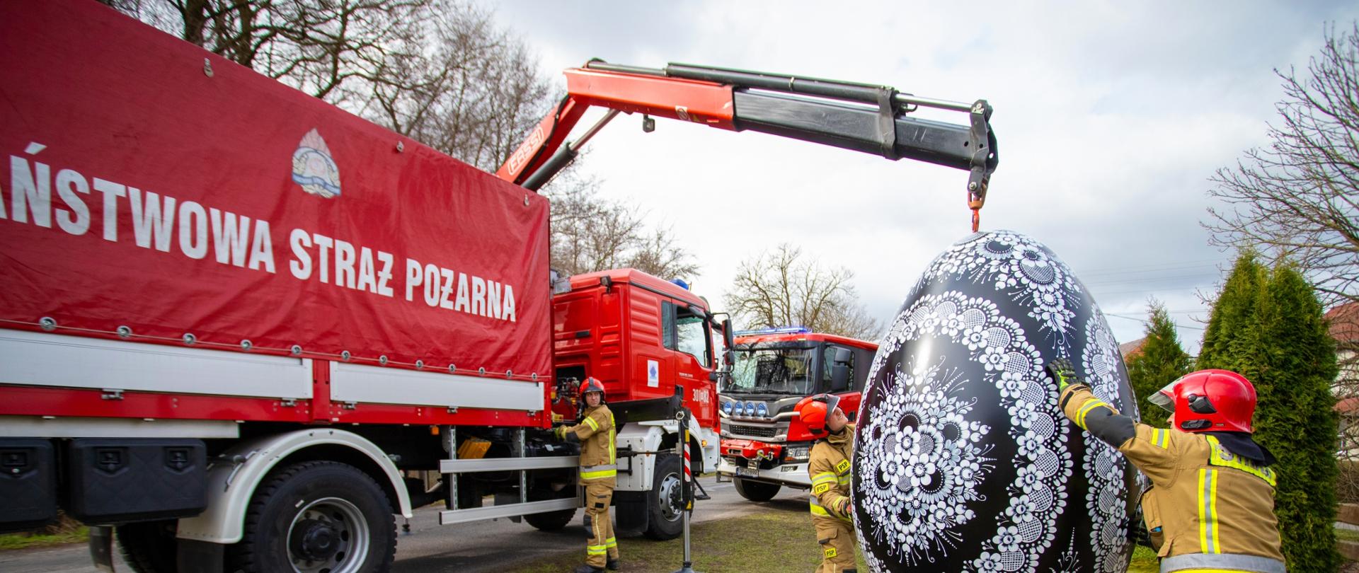 Strażacy Państwowej Straży Pożarnej z Opola ustawiają dużą, ozdobną pisankę wielkanocną przy użyciu samochodu z dźwigiem. W tle stoją wozy strażackie, a cała scena odbywa się na trawiastym terenie. Na grafice widnieją życzenia wielkanocne podpisane przez Opolskiego Komendanta Wojewódzkiego PSP, st. bryg. Arkadiusza Kuśmierskiego.
Z okazji Świąt Wielkanocnych, niosących nadzieję i odrodzenie życia, pragnę złożyć najserdeczniejsze życzenia.
Niech ten radosny, świąteczny czas, spędzony bez pośpiechu, troski i zmartwień, w zdrowiu oraz rodzinnej atmosferze, będzie źródłem nadziei, siły oraz wiary.
W imieniu strażaków województwa opolskiego
Opolski Komendant Wojewódzki Państwowej Straży Pożarnej
st. bryg. Arkadiusz Kuśmierski