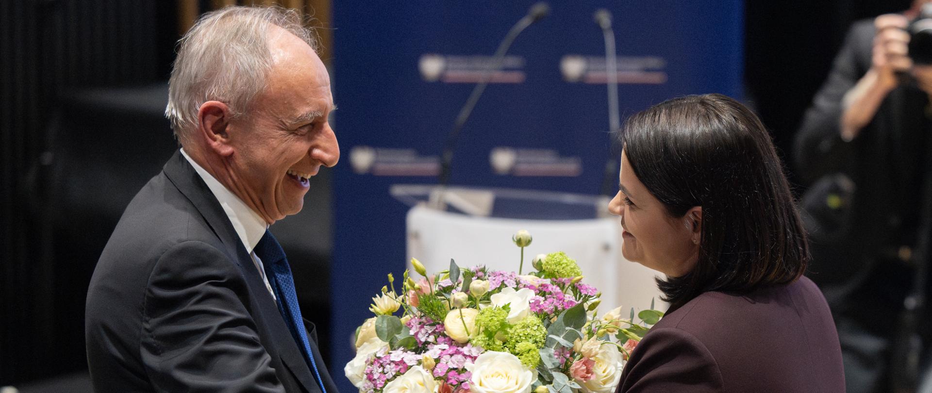 Na zdjęciu dwie osoby - jedna wręcza kwiaty drugiej. W tle scena.