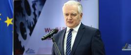 Wicepremier, minister rozwoju, pracy i technologii Jarosław Gowin stojący przy mównicy, z tyłu za jego placami flaga UE oraz ekran z napisem Wyniki Białej Księgi Rozwoju Przemysłu