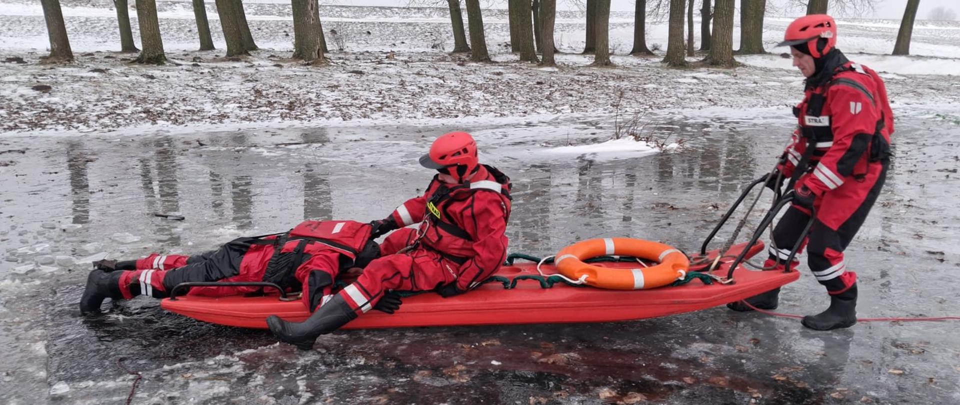 Strażacy z JRG Koło podczas ćwiczeń z ratownictwa lodowego. Jeden strażak leży na saniach lodowych. Sanie lodowe prowadzone są przez drugiego ratownika. Ratownicy oraz sanie zabezpieczeni są linami. Zdjęcie wykonane na pokrywie lodowej, widać wyraźnie zalegający śnieg.
