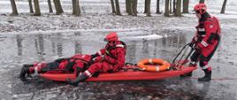 Strażacy z JRG Koło podczas ćwiczeń z ratownictwa lodowego. Jeden strażak leży na saniach lodowych. Sanie lodowe prowadzone są przez drugiego ratownika. Ratownicy oraz sanie zabezpieczeni są linami. Zdjęcie wykonane na pokrywie lodowej, widać wyraźnie zalegający śnieg.
