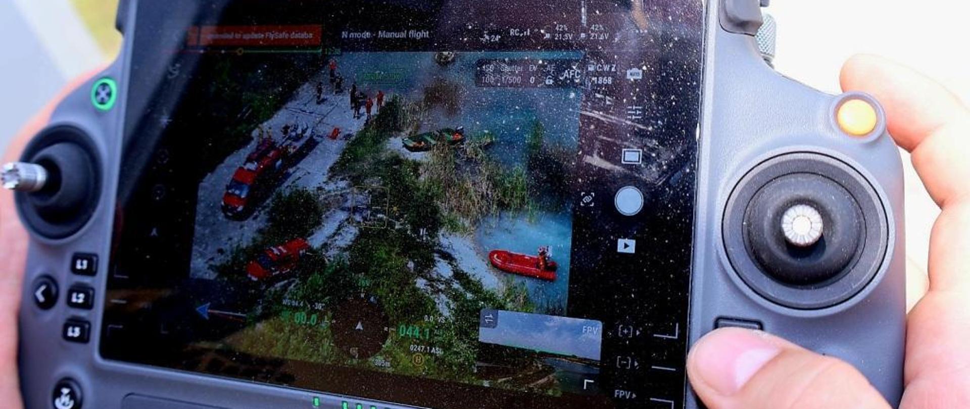 Zdjęcie wykonane podczas ćwiczeń na terenie zalanej przez wodę kopalni piachu w miejscowości Wierzbica. Na zdjęciu widać wyświetlacz urządzenia sterowniczego do obsługi drona. Na ekranie wyświetlacza widać pojazdy ratowniczo – gaśnicze w kolorze czerwonym ze srebrnymi i białymi elementami zabudowy oraz strażaków ze specjalistycznej grupy ratownictwa wodno – nurkowego. Strażacy przygotowują się do zwodowania łodzi na wodę tworzącą jezioro w zalanym nieczynnym wyrobisku kopalni. 