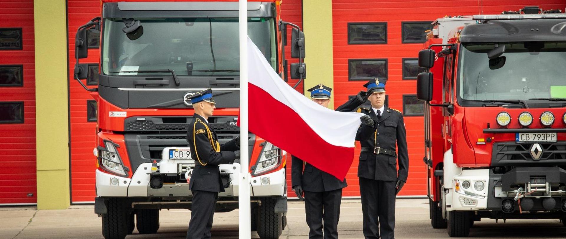 Trzech strażaków stoi przy maszcie flagowym. Jeden zaczyna podnosić flagę Polski w górę na maszt. Dwóch strażaków salutuje. Za masztem stoją samochody strażackie,