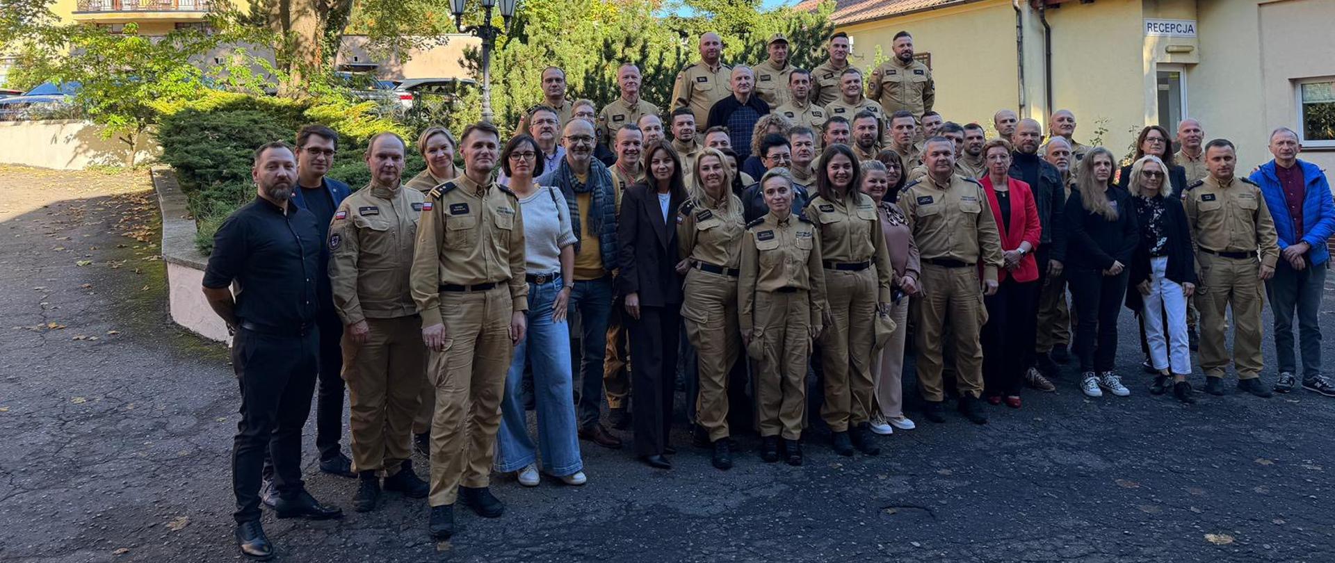 Na zdjęciu widać dużą grupę około osób stojących na zewnątrz, przed jasnym budynkiem z napisem „Recepcja”. Większość osób ma na sobie piaskowe mundury, niektórzy ubrani są w cywilne stroje – marynarki, kurtki, swetry czy jeansy. Grupa stoi w kilku rzędach, część osób uśmiecha się do aparatu. W tle rosną wysokie drzewa z zielonymi i żółtymi liśćmi. Nad grupą widać niebieskie, bezchmurne niebo.