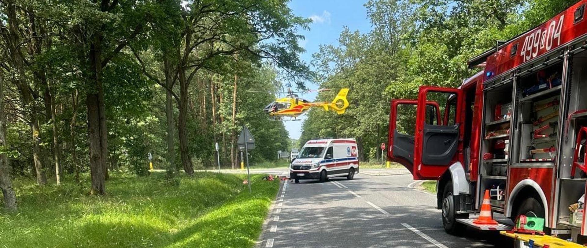 Na zdjęciu znajduje się pojazd straży pożarnej, karetka pogotowia oraz śmigłowiec LPR podchodzący do lądowania
