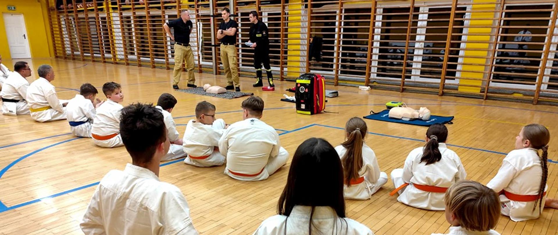 Zdjęcie przedstawia sekcje karate Kyokushin na sali gimnastycznej. Zawodnicy siedzą na podłodze a przed nimi stoją strażacy PSP, na ziemi znajdują się fantomy i torba R1.