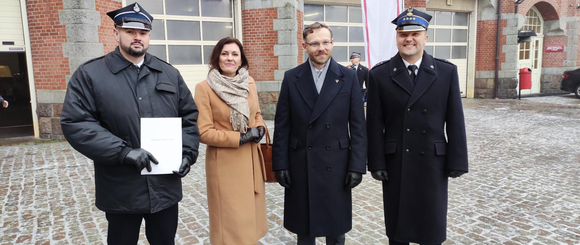 Na zdjęciu widoczna w tle murowana z czerwonej cegły zabytkowa ściana budynku strażnicy z bramami garażowymi. Na pierwszym planie stoją na pokrytym brukiem placu, patrzący na wprost osoby, w kolejności od lewej: mężczyzna z ciemnymi wąsami i brodą, w czarnych butach, spodniach i kurtce mundurowej, z rogatywką na głowie, trzymający w czarnych rękawiczkach dokument; kobieta w wysokich czarnych kozakach, beżowym płaszczu i jasnobrązowym szaliku na szyi; mężczyzna w czarnych butach, szarych spodniach, z szalikiem wystającym spod granatowego długiego płaszcza; mężczyzna z rogatywką na głowie, w czarnych butach, spodniach i granatowym długim płaszczu z pagonami, spod którego widoczna biała koszula z czarnym krawatem.