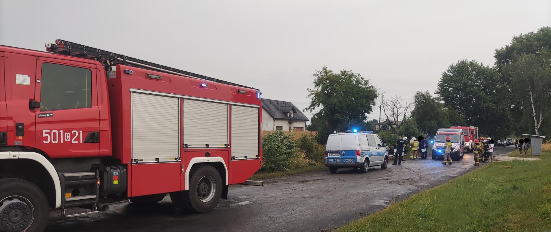 Zdjęcie przedstawia jezdnie drogi powiatowej na której znajdują się pojazdy służb ratowniczych (dwa wozy straży pożarnej, jeden radiowóz policji, pogotowie ratunkowe) w trakcie zdarzenia drogowego po dachowaniu pojazdu osobowego.