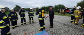Zdjęcie przedstawia ubierającego się strażaka w aparat ochrony układu oddechowego. Z lewej strony czterech obserwujących strażaków. Z prawej strony funkcjonariusz PSP nadzorujący wykonanie zadania. Na kostce przed strażakami leży pilarka do drewna.