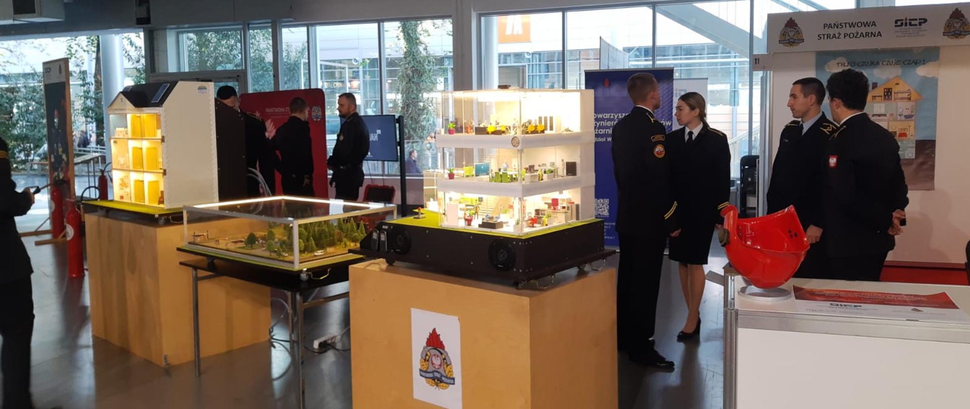 grupa strażaków stoi na stoisku targowym, przed nimi stoją trzy symulatory zagrożeń