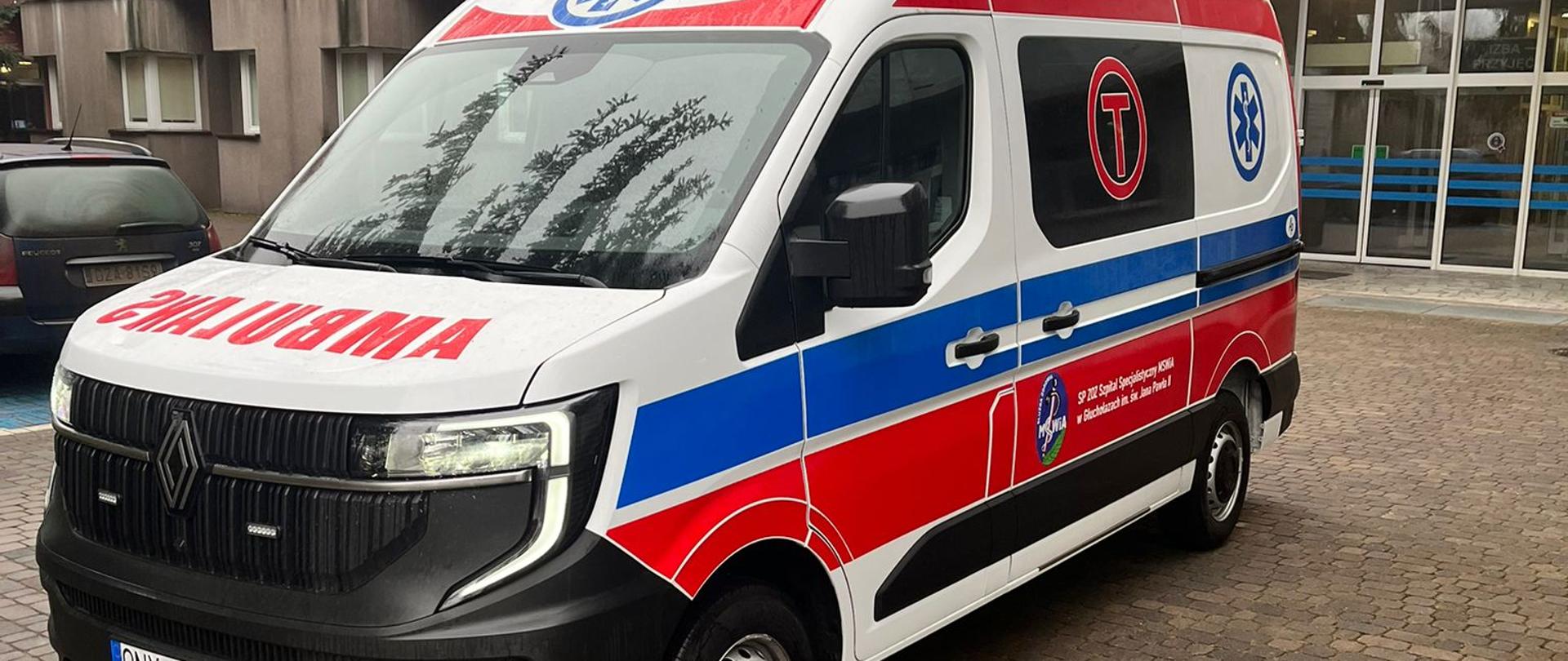 Na zdjęciu widoczna jest nowoczesna karetka pogotowia ratunkowego, zaparkowana na brukowanym podjeździe przed budynkiem. Karetka jest biała z charakterystycznymi czerwono-niebieskimi pasami po bokach, symbolami gwiazdy życia oraz napisem "AMBULANS" z przodu i po bokach. Na dachu pojazdu znajdują się niebieskie światła sygnalizacyjne. Na drzwiach bocznych widoczny jest herb oraz tekst w języku polskim, w tym "SP ZOZ Szpital Specjalistyczny MSWiA w Głuchołazach im. Jana Pawła II". W tle znajduje się budynek szpitalny lub placówki medycznej z szarymi i czerwonymi elewacjami oraz dużymi oknami. Podłoże jest mokre, co sugeruje niedawny deszcz.