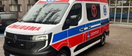Na zdjęciu widoczna jest nowoczesna karetka pogotowia ratunkowego, zaparkowana na brukowanym podjeździe przed budynkiem. Karetka jest biała z charakterystycznymi czerwono-niebieskimi pasami po bokach, symbolami gwiazdy życia oraz napisem "AMBULANS" z przodu i po bokach. Na dachu pojazdu znajdują się niebieskie światła sygnalizacyjne. Na drzwiach bocznych widoczny jest herb oraz tekst w języku polskim, w tym "SP ZOZ Szpital Specjalistyczny MSWiA w Głuchołazach im. Jana Pawła II". W tle znajduje się budynek szpitalny lub placówki medycznej z szarymi i czerwonymi elewacjami oraz dużymi oknami. Podłoże jest mokre, co sugeruje niedawny deszcz.