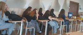 Zdjęcie przedstawia grupę młodzieży siedzącą w ławkach szkolnych na tle nietypowej fototapety z gęsiami.Opis pierwszego planuW dolnej części kadru widoczna jest klasa lekcyjna. Wzdłuż rzędu drewnianych brązowych biurek z metalowymi stelażami siedzi tyłem lub bokiem do obiektywu około siedmiu osób, głównie dziewcząt z długimi włosami. Są ubrani w codzienne stroje: jeansy, dresy i ciemne koszulki. Część osób opiera się o blaty, rozmawia lub patrzy w stronę niewidocznego dla widza przodu sali. Na krzesłach i ławkach leżą plecaki.Opis tła i otoczeniaCałą ścianę za uczniami zajmuje ogromna, jasna fototapeta przedstawiająca cztery białe gęsi z pomarańczowymi dziobami. Ptaki są ukazane od piersi w górę i wydają się „zaglądać” do sali lekcyjnej. W prawym górnym rogu, pod sufitem, podwieszona jest metalowa rura z czerwono-pomarańczowymi elementami automatycznego systemu karmienia lub pojenia drobiu. Po prawej stronie widoczne są zamknięte drewniane drzwi, a obok nich na stoliku stoją plastikowe kanistry i butelki z produktami weterynaryjnymi lub paszowymi.Kompozycja i oświetlenieZdjęcie zostało zrobione z boku sali, z wysokości oczu siedzących osób. Pomieszczenie jest jasno oświetlone sztucznym, ciepłym światłem sufitowym. Dominują odcienie bieli, szarości, brązu oraz wyraziste pomarańczowe akcenty dziobów i elementów wyposażenia rolniczego. Kadrowanie łączy codzienny widok lekcji z surrealistycznym motywem hodowlanym na ścianie.