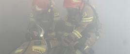 zadymione pomieszczenie, ledwo widać dwóch funkcjonariuszy PSP w pełni ubranych w środki ochrony indywidualnej, którzy po ziemi ciągną druha OSP w pełnym umundurowaniu z podpiętym aparatem ochrony układu oddechowego.