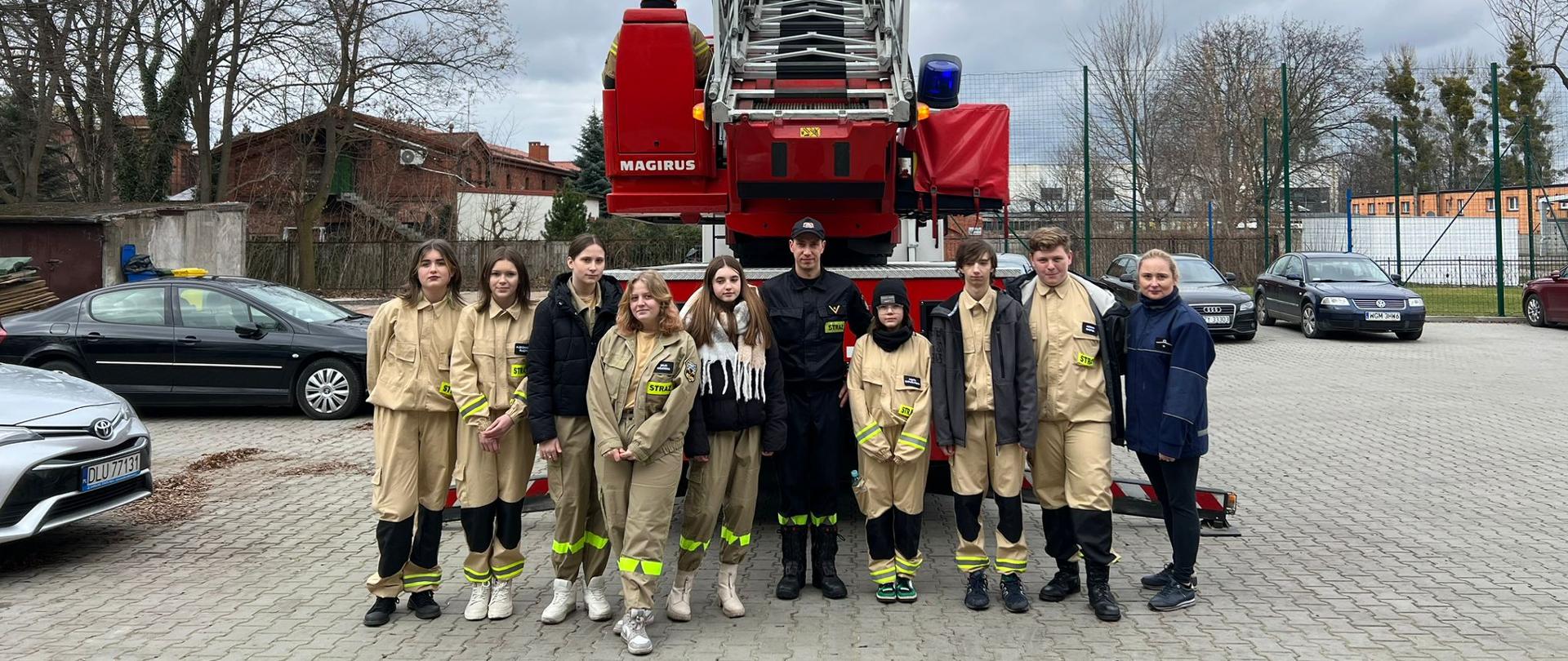 Grupowe zdjęcie uczestników wycieczki wraz ze strażakiem z KP PSP w Żyrardowie za tle rozłożonej w górę drabiny z wozu strażackiego