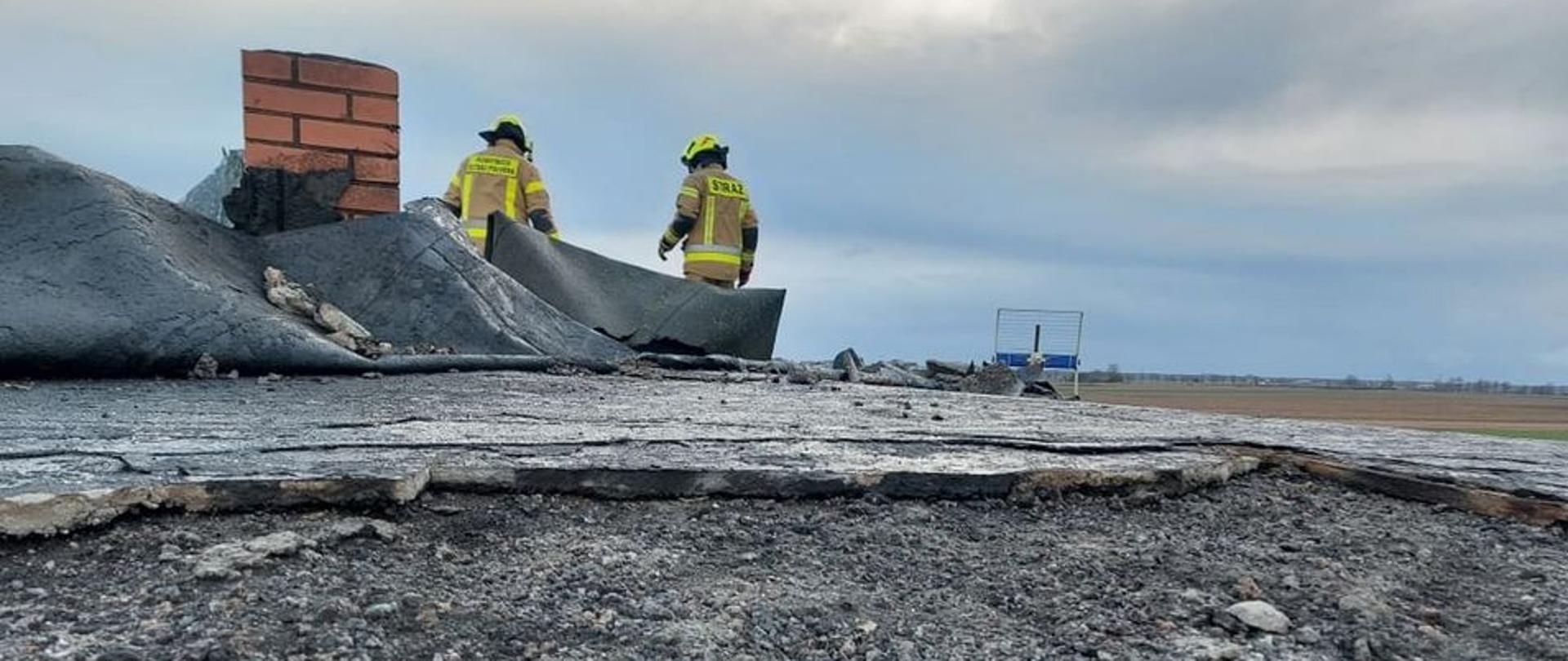 Pochmurno. Zerwane poszycie dachu. Duży fragment czarnej papy odsłania betonowy strop i leży przy kominie pogięty. Komin z czerwonej cegły. Przy nim dwóch strażaków w jasnych mundurach.