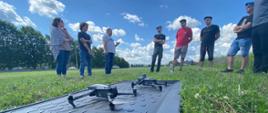 Grupa jedenastu osób uczestniczy w szkoleniu terenowym na polu doświadczalnym. Uczestnicy stoją w półkolu i słuchają prowadzącego. Na trawie przed nimi znajdują się dwa drony, aparatura sterująca oraz walizki transportowe ze sprzętem. W tle widoczne są poletka doświadczalne, zabudowa jednorodzinna oraz częściowo zachmurzone niebo. Zdjęcie dokumentuje praktyczną część szkolenia z wykorzystania bezzałogowych statków powietrznych do monitorowania upraw.