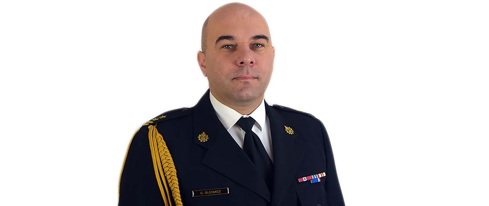 Zdjęcie wykonane na białym tle przedstawia profil komendanta powiatowego PSP w Sanoku bryg. Grzegorza Oleniacza. Komendant ubrany jest w granatowy mundur wyjściowy PSP ze złotym sznurem.