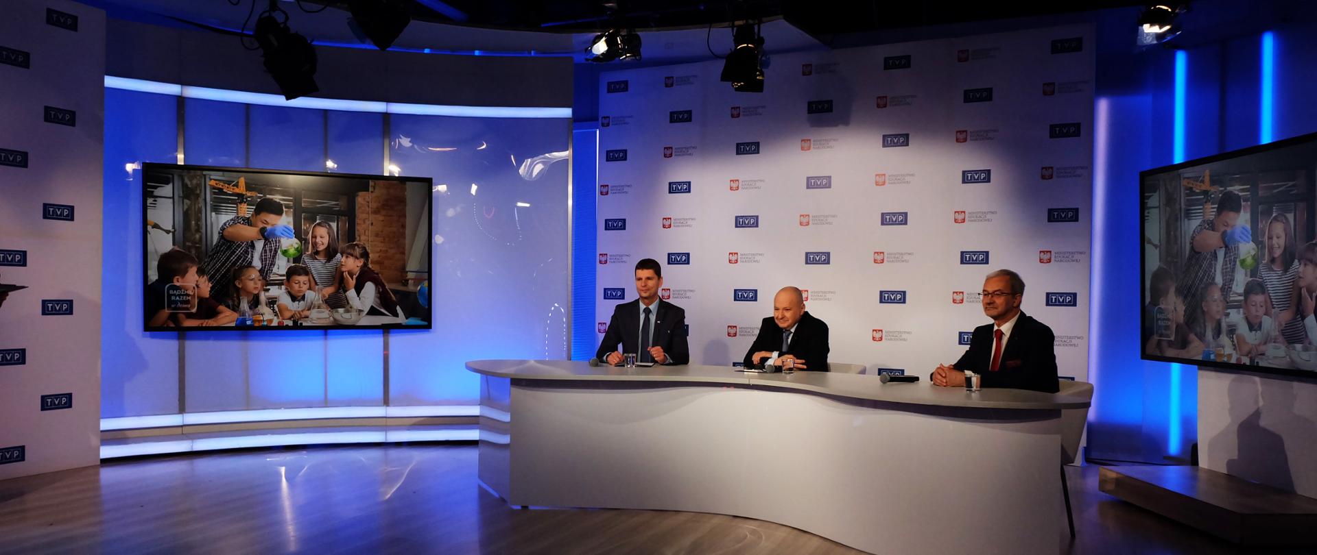 Studio telewizyjne. Po obu stronach stoją monitory ekranowe. W środku za stołem siedzi minister edukacji, prezes TVP i szef PGNiG