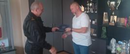 Komendant Powiatowy PSP w Rypinie starszy brygadier Grzegorz Łydkowski wręcza list gratulacyjny z okazji jubileuszu trzydziestolecia pracy starszemu technikowi Marcinowi Dłużniewskiemu. Stoją oni na wprost, podają sobie dłonie, a Komendant Powiatowy PSP w Rypinie przekazuje Panu Marcinowi Dłużniewskiemu list gratulacyjny. Znajdują się Oni w gabinecie Komendanta. W tle widać szafki i gabloty na puchary.