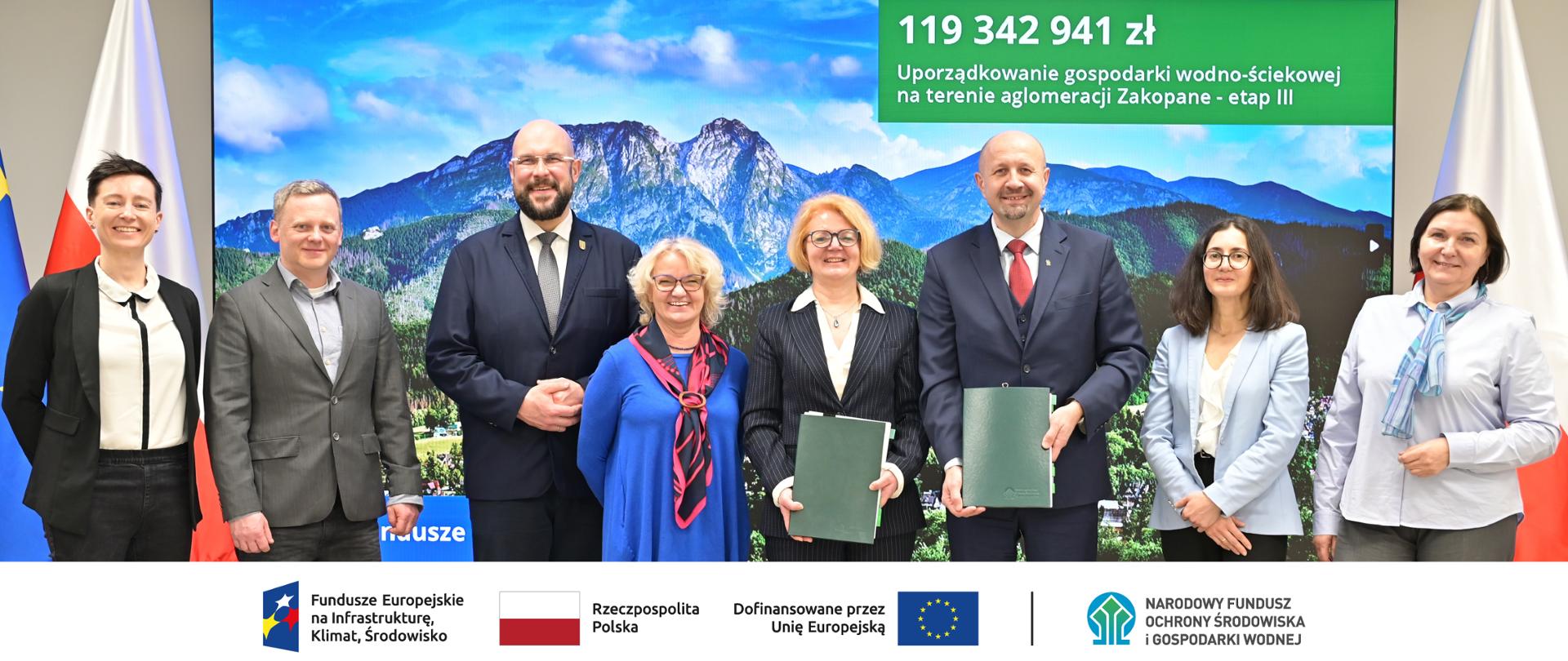Podpisanie umowy. Grupa 8 osób pozujących do zdjęcia, wśród nich: Robert Gajda - zastępca prezesa zarządu NFOŚiGW, Stanisława Czubernat - prezes zarządu SEWiK, Marcin Rafacz - prokurent SEWiK, Łukasz Filipowicz - burmistrz Zakopanego. Nad nimi widnieje napis: „119 342 941 zł – Uporządkowanie gospodarki wodno-ściekowej na terenie aglomeracji Zakopane – etap III”. Na dole grafiki znajdują się logotypy: Fundusze Europejskie na Infrastrukturę, Klimat, Środowisko, Rzeczpospolita Polska, Dofinansowane przez Unię Europejską oraz Narodowy Fundusz Ochrony Środowiska i Gospodarki Wodnej.