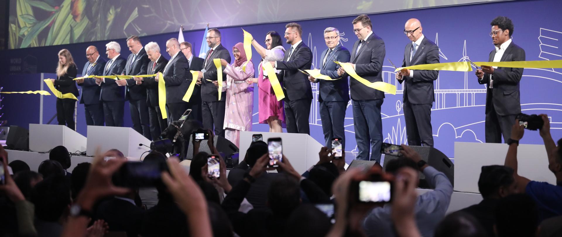 Na scenie grupa osób przecina wstęgę. Przed nimi dziennikarze z aparatami.