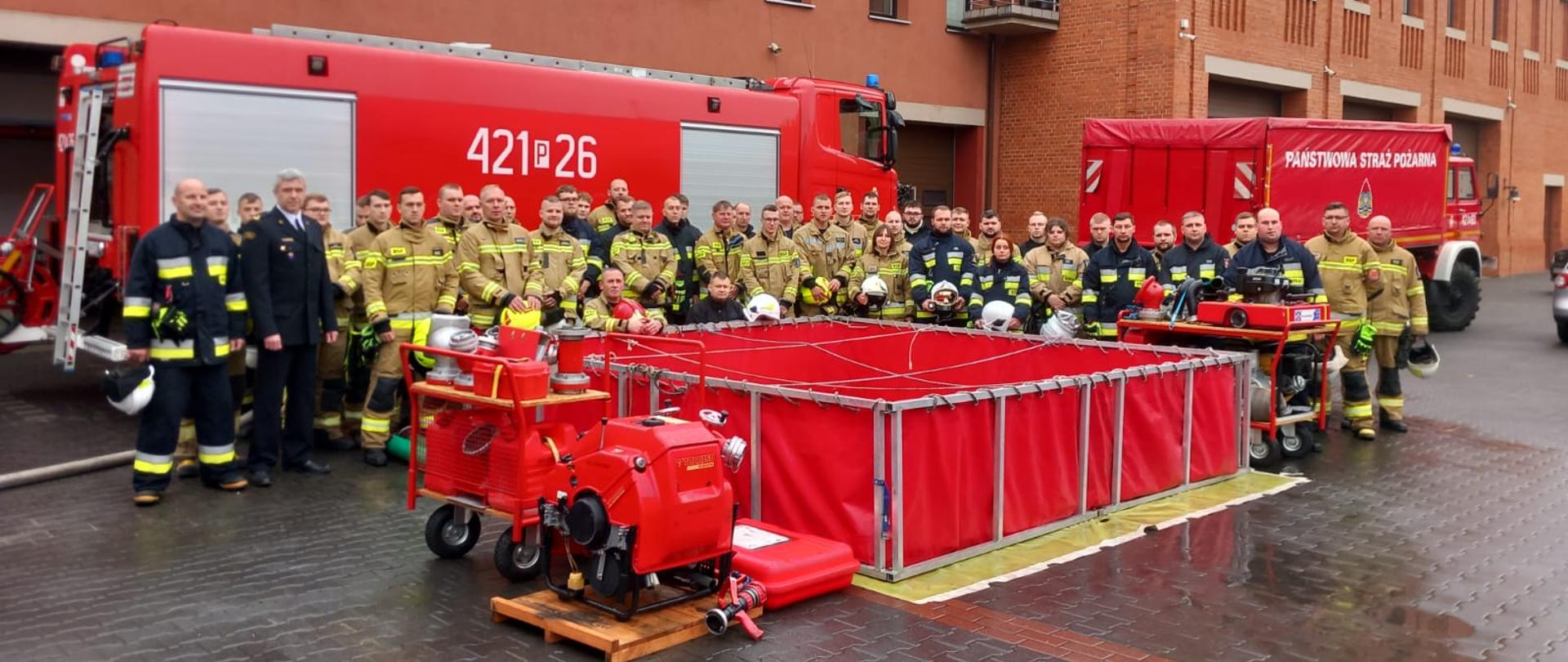 Na grupowym zdjęciu widać osoby biorących udział w szkoleniu. Stoją oni na tle czerwonego samochodu pożarniczego. Przed całą grupą rozłożony jest zbiornik a obok stoi motopompa