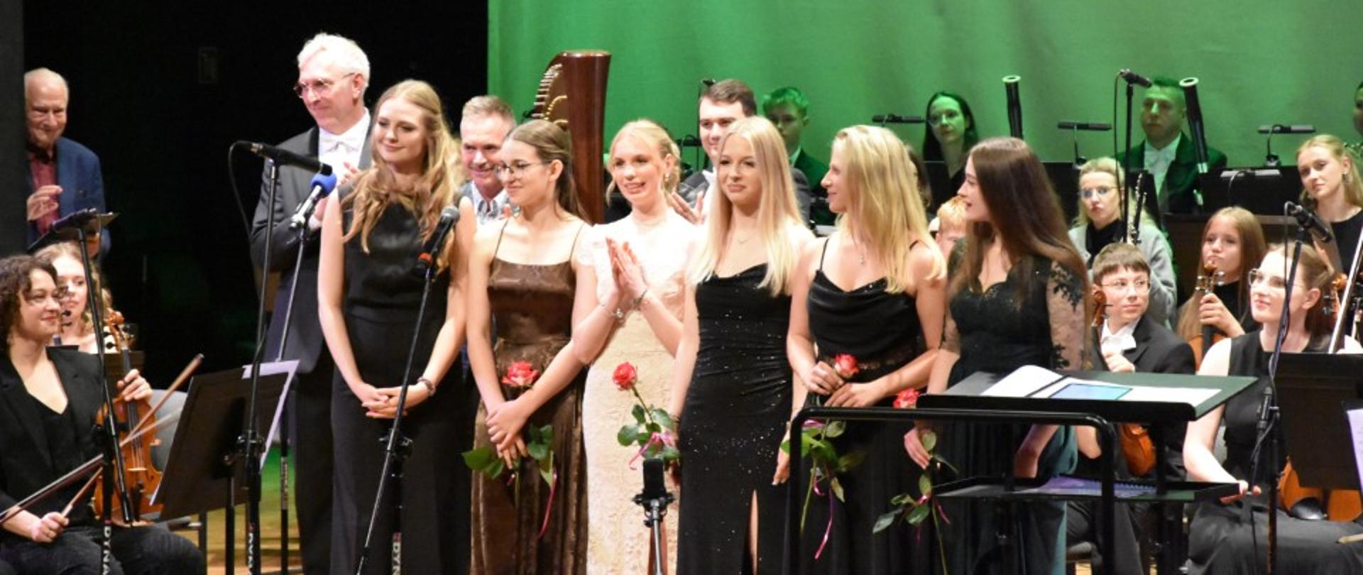Zdjęcie kolorowe. Grupa artystów stojących na scenie podczas koncertu lub uroczystej gali. W pierwszym rzędzie stoi sześć młodych kobiet w eleganckich sukniach wieczorowych – przeważają kreacje w kolorze czarnym, ciemnobrązowym oraz jedna długa, jasna sukienka. Niektóre z nich trzymają w dłoniach pojedyncze, czerwone róże. Za nimi stoją mężczyźni w garniturach i garniturowych koszulach oraz inni członkowie orkiestry, w tym osoby z instrumentami takimi jak skrzypce. Po lewej stronie, w tle, stoi mężczyzna w okularach i ciemnej marynarce. Po prawej stronie siedzą muzycy z instrumentami. W tle znajduje się duża, gładka, zielona kurtyna, a na scenie ustawione są mikrofony na statywach oraz pulpity na nuty.