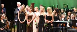 Zdjęcie kolorowe. Grupa artystów stojących na scenie podczas koncertu lub uroczystej gali. W pierwszym rzędzie stoi sześć młodych kobiet w eleganckich sukniach wieczorowych – przeważają kreacje w kolorze czarnym, ciemnobrązowym oraz jedna długa, jasna sukienka. Niektóre z nich trzymają w dłoniach pojedyncze, czerwone róże. Za nimi stoją mężczyźni w garniturach i garniturowych koszulach oraz inni członkowie orkiestry, w tym osoby z instrumentami takimi jak skrzypce. Po lewej stronie, w tle, stoi mężczyzna w okularach i ciemnej marynarce. Po prawej stronie siedzą muzycy z instrumentami. W tle znajduje się duża, gładka, zielona kurtyna, a na scenie ustawione są mikrofony na statywach oraz pulpity na nuty.