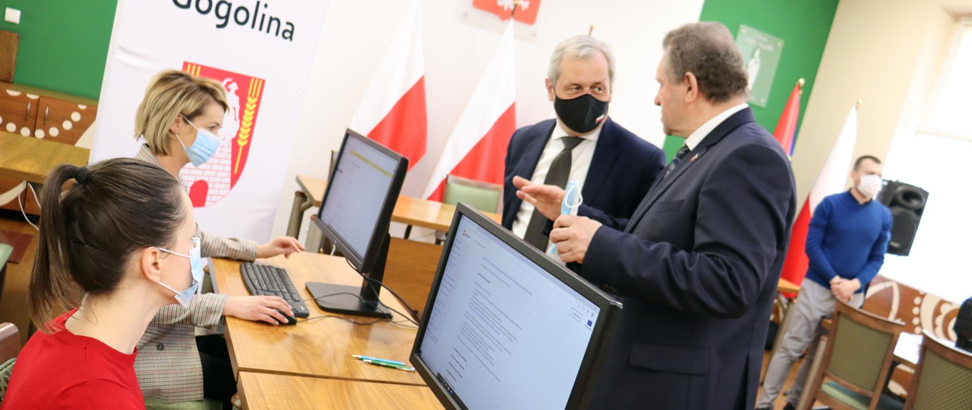 Wojewoda opolski odwiedza punkt rejestracji nr pesel w UG Gogolin. Na zdjęciu po lewej stronie dwie pracownice urzędu, siedzące przy biurku. Przed nimi komputer. Na przeciw nich, stoją wojewoda opolski i burmistrz gminy. Spoglądają na siebie, rozmawiają. W tle biało-czerwone flagi, stół oraz pozostali uczestnicy spotkania